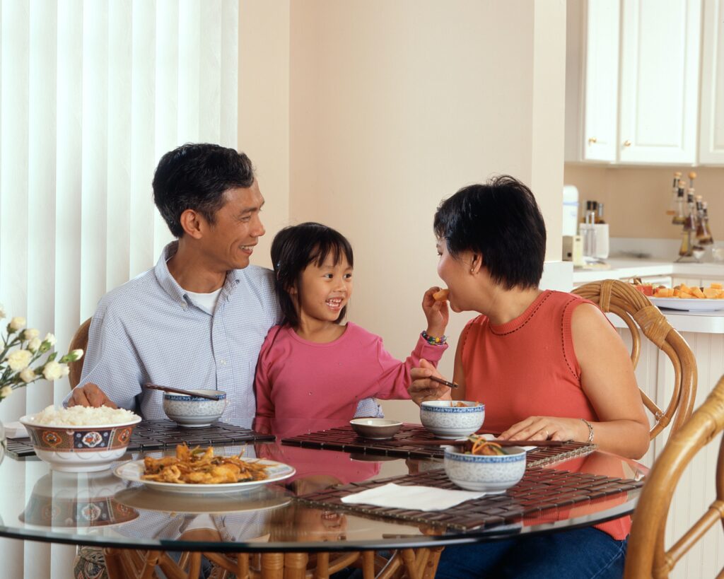 Parenting Adalah Tentang Membantu Anak anak Belajar dan Tumbuh ifmama – An Asian family an adult male and female are seated around a table eating a meal with a young female standing in between the adults